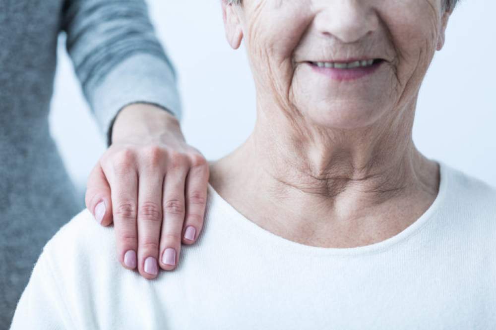 Mulher idosa de frente com uma mão ao ombro de uma cuidadora