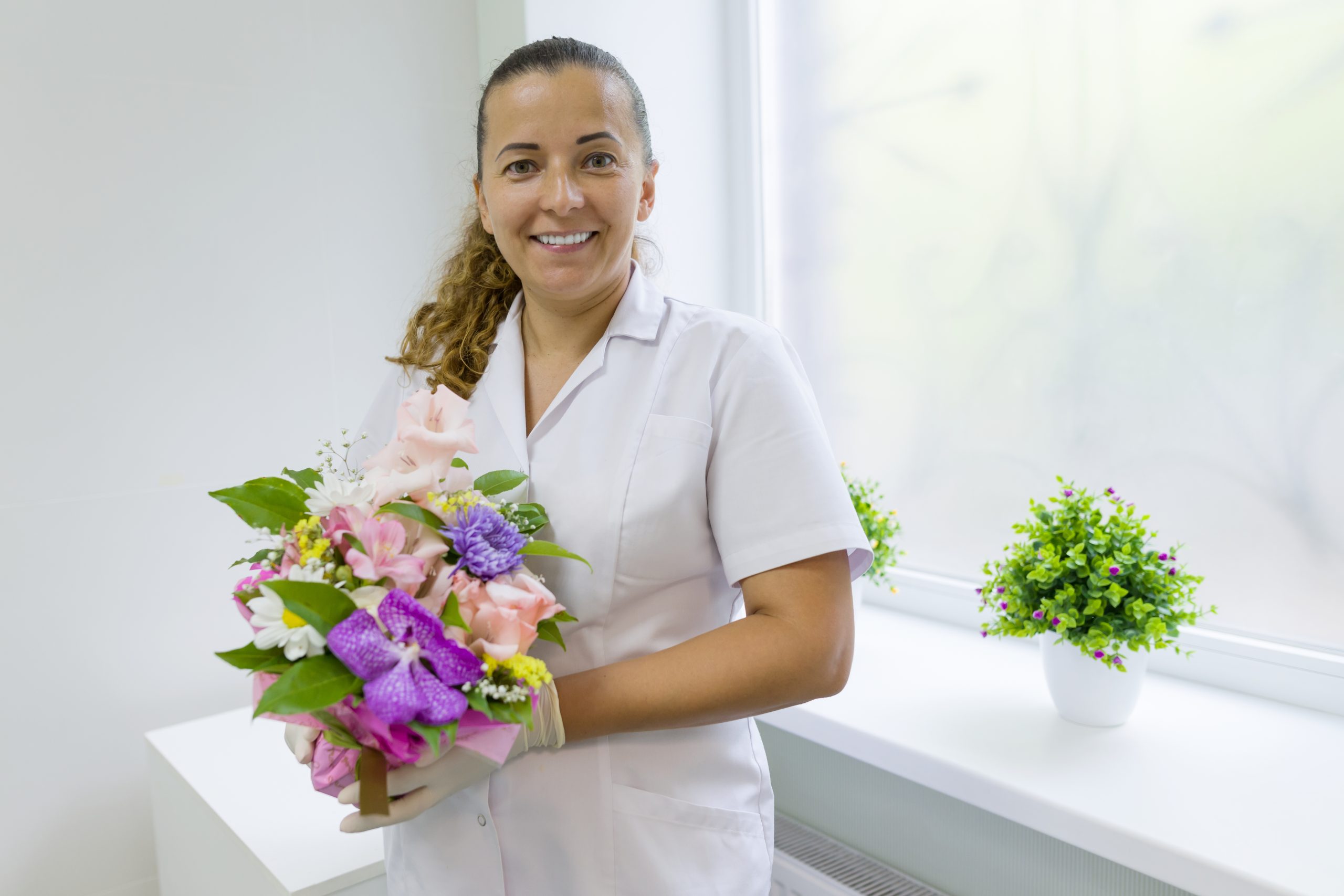 Enfermeira com buquê de flores, sorrindo perto da janela do hospital. Dia Nacional do Médico.